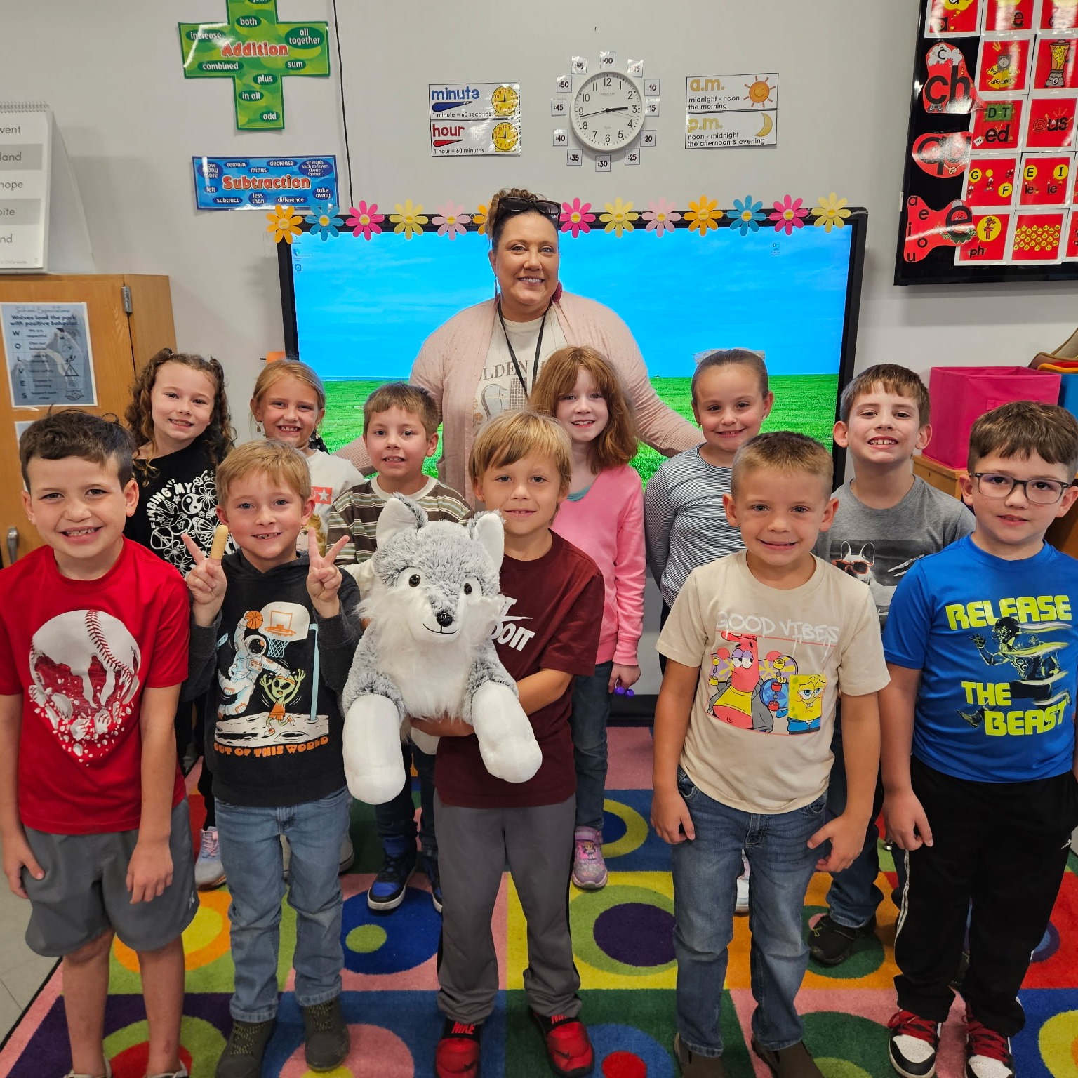 Classroom with children and teacher; some children hold a white stuffed animal. Brightly colored rug.