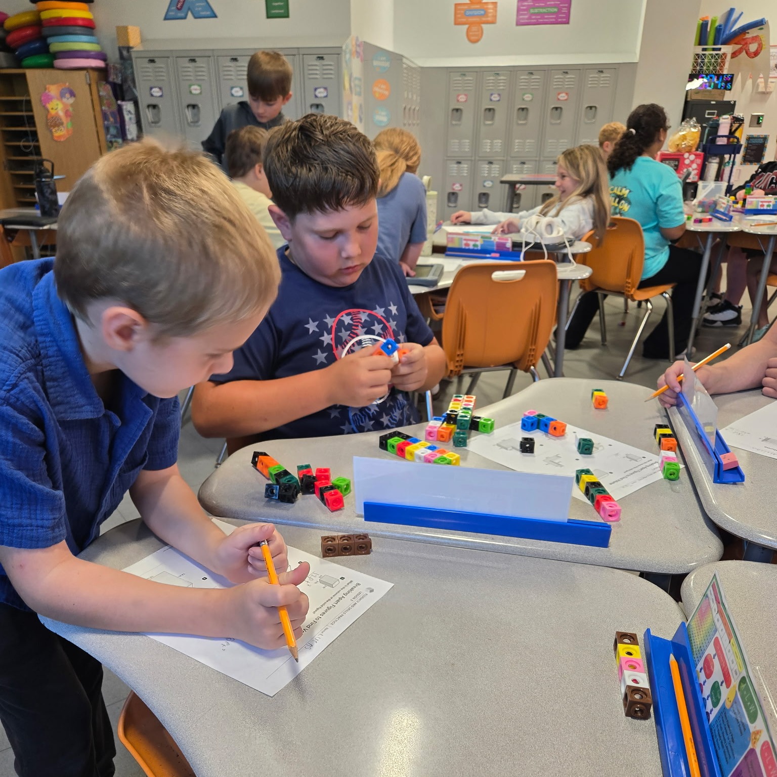 A classroom with students at desks. Two boys work on a puzzle with small colored cubes.