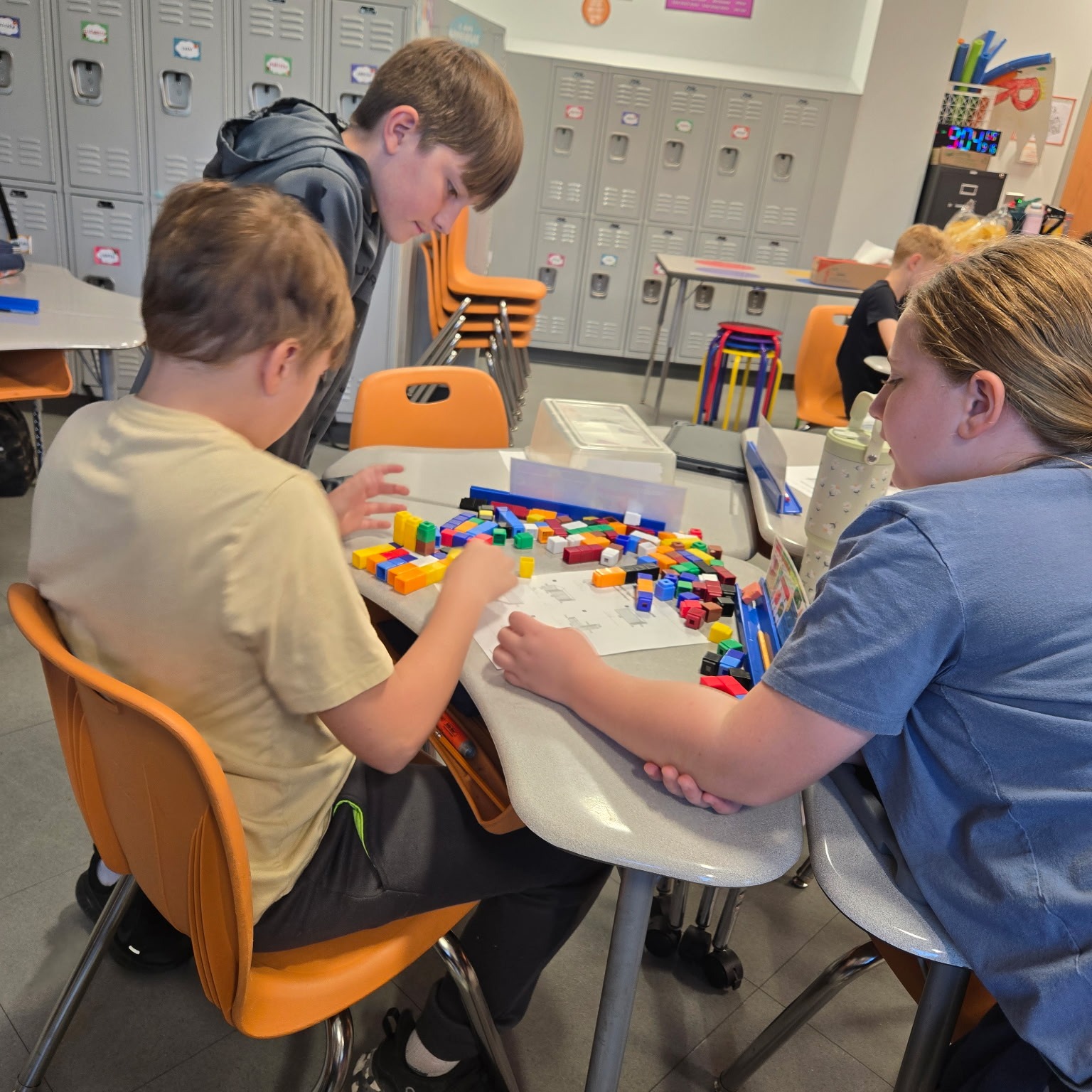 Three children at desks in a classroom, two playing with multicolored blocks on a table.