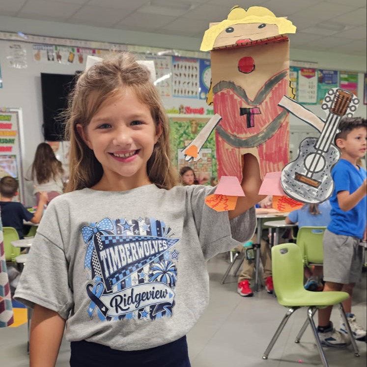 Girl holds a puppet of a figure holding a guitar in a classroom with other students.