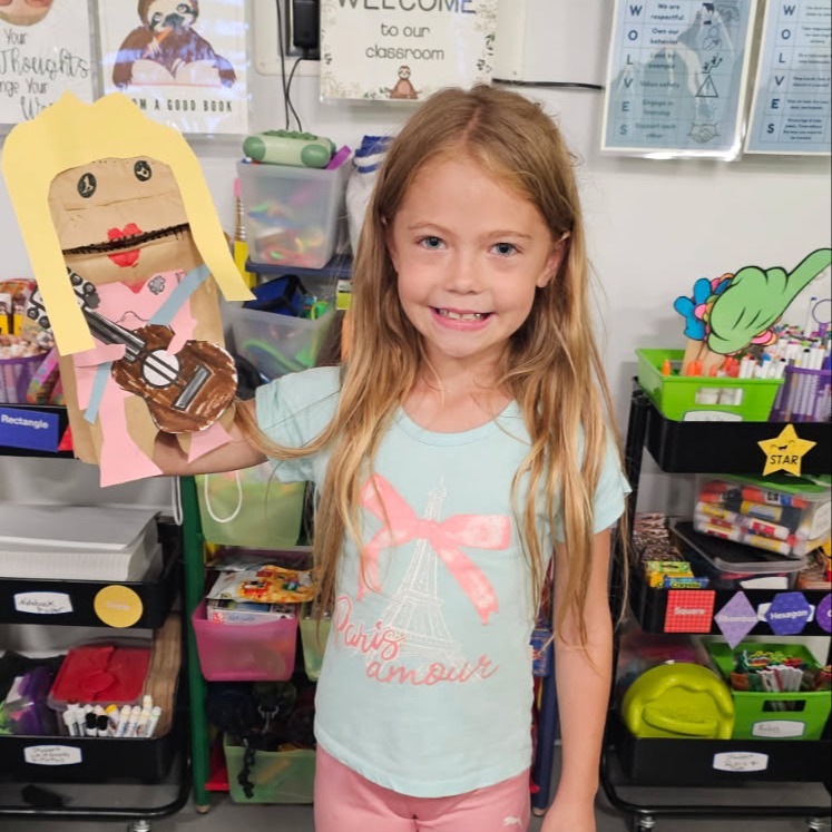 A girl holds a paper cutout of a person holding a guitar while standing in a classroom.