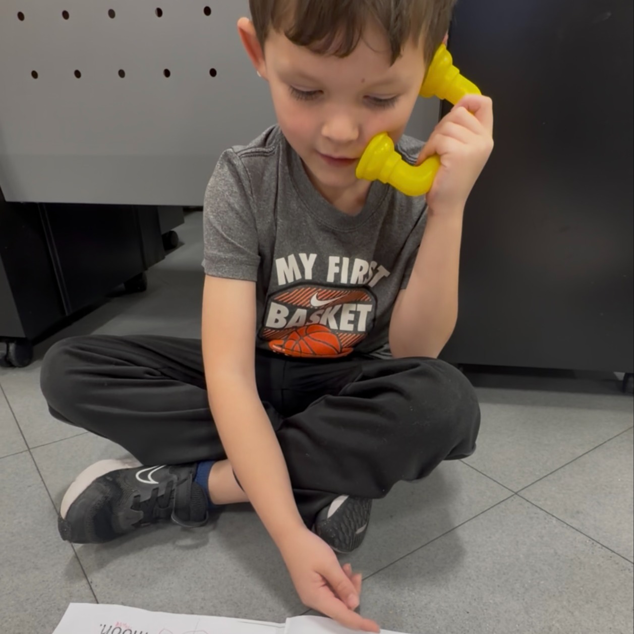 Young boy wearing black pants sitting on floor holding up two yellow objects and reading from an open book.