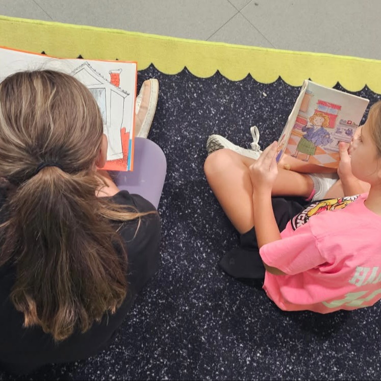 Two young girls sitting on a floor reading books, one with a ponytail, the other with a pigtail.