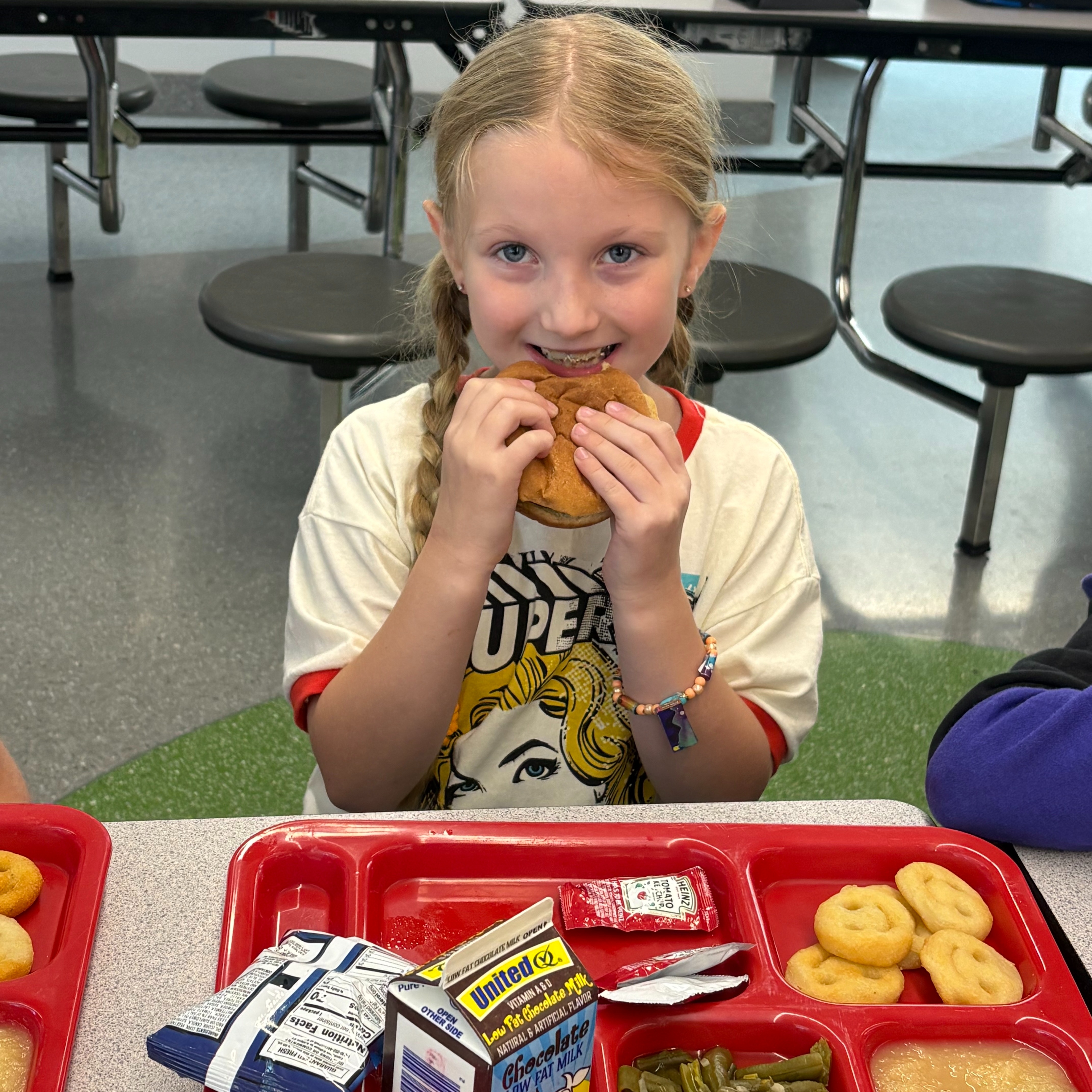 Girl in white shirt sitting at cafeteria table eating sandwich, tray with food and drinks in front.