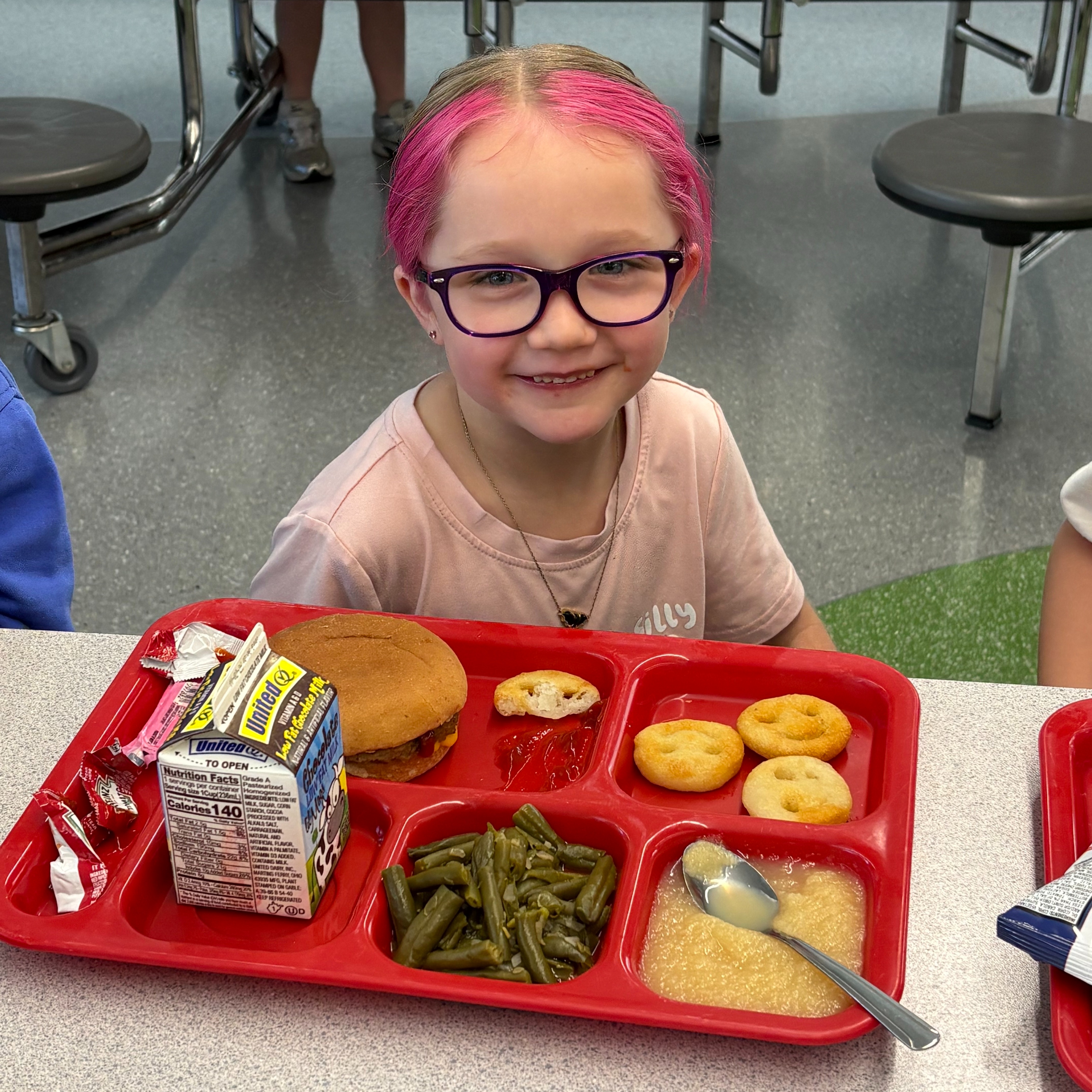 A young girl in pink sits at a cafeteria table with a tray of food.