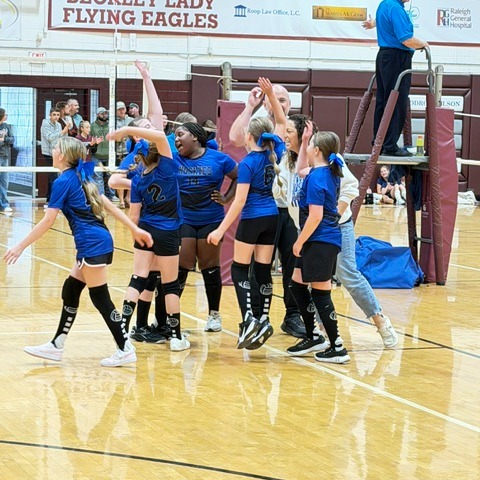 Several girls in blue uniforms celebrating on a court with a referee watching from above.