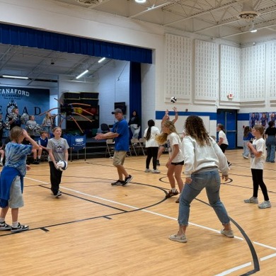 Several children playing with stick-like objects on an indoor court with a basketball hoop and seated spectators.