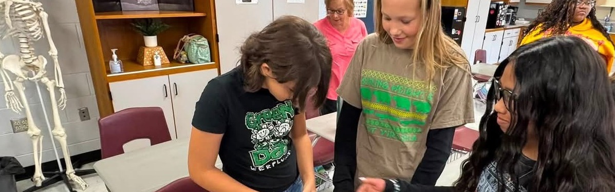 Four students examine a dummy torso with a bandage. A skeleton stands on a shelf in the background.