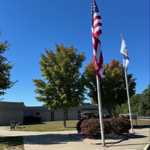 A flagpole with American and state flags in front of a school building. Trees and bushes are in the foreground.