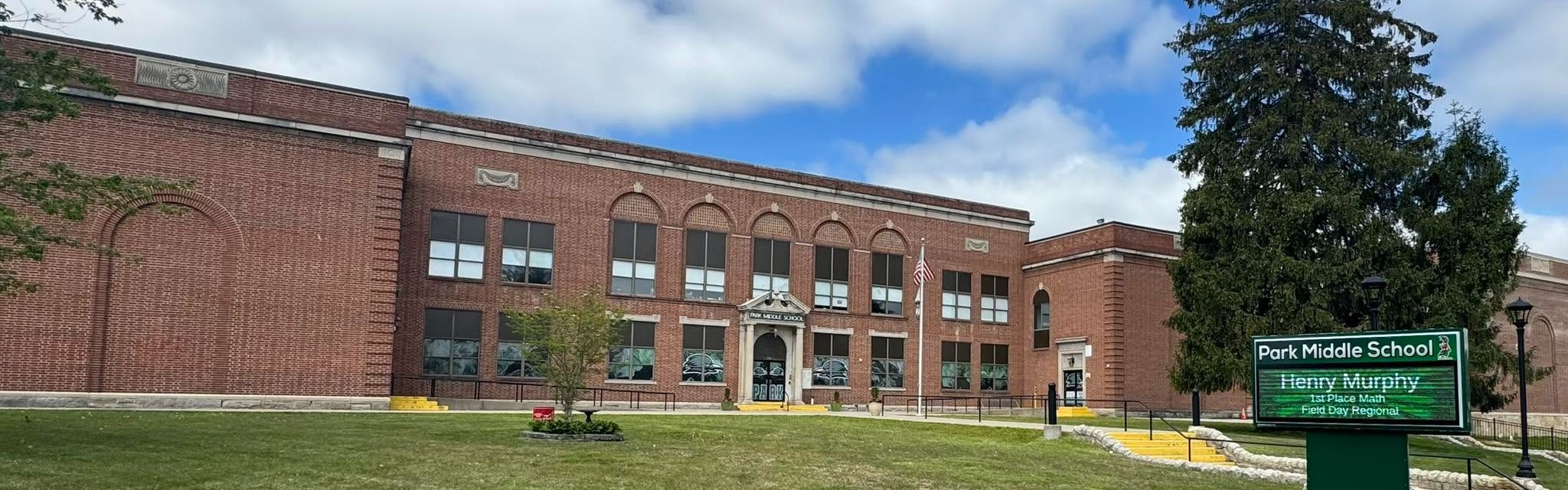 Large brick school building with many windows and a green sign in front. Green lawn and trees.