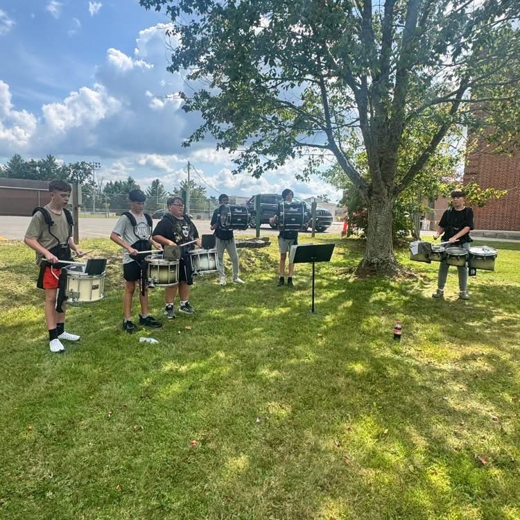Six children play drums outside on a grassy field. A tree is nearby. A building and parked vehicles are in the background.