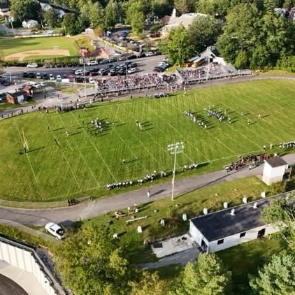 Aerial view of a football field with people on the field, bleachers, and cars parked along the road.