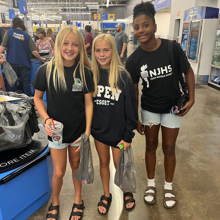 Three girls stand in a store aisle. Two wear black shirts; one has "PEW" and the other has "NJHS."