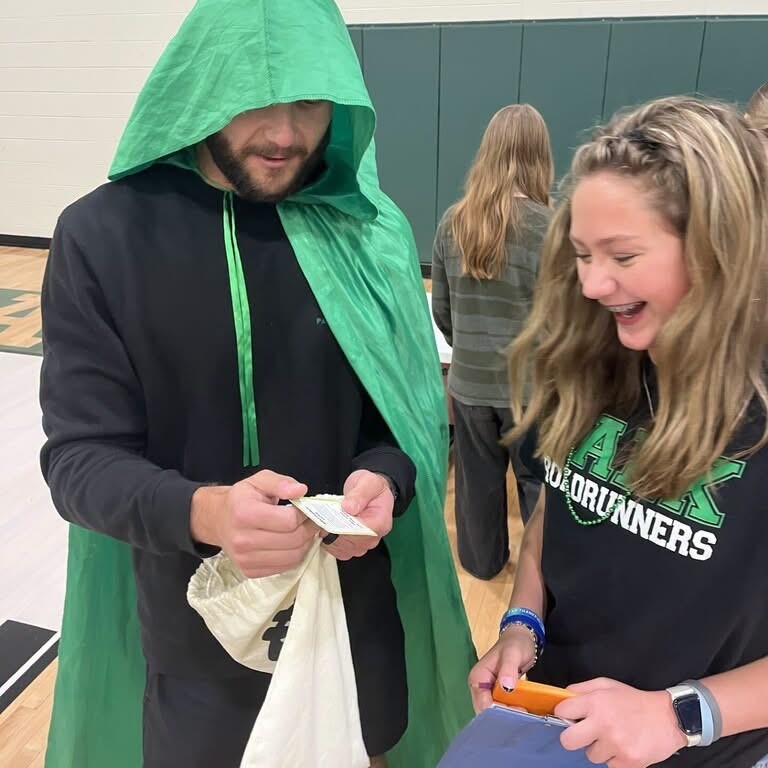 Person in green cape holding paper. Person in black shirt holding blue folder, smiling. Two women standing in background, one wearing gray top.