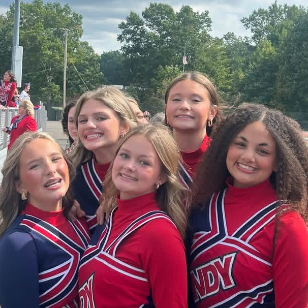 Five girls in red and blue uniforms smile for photo in front of bleachers with trees.