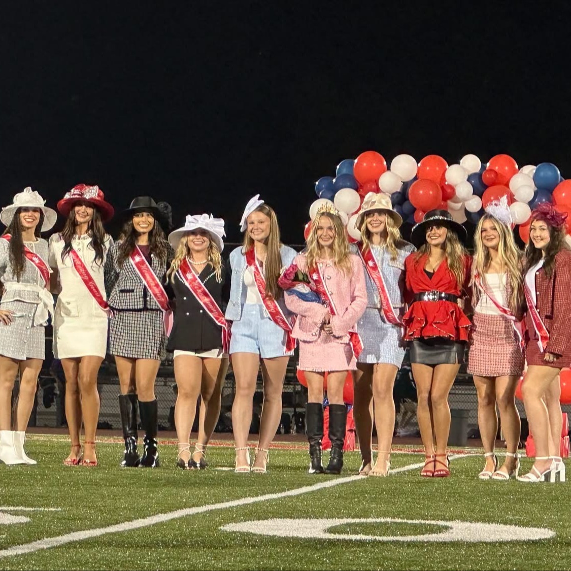 A group of women in hats and sashes standing on a football field with balloons