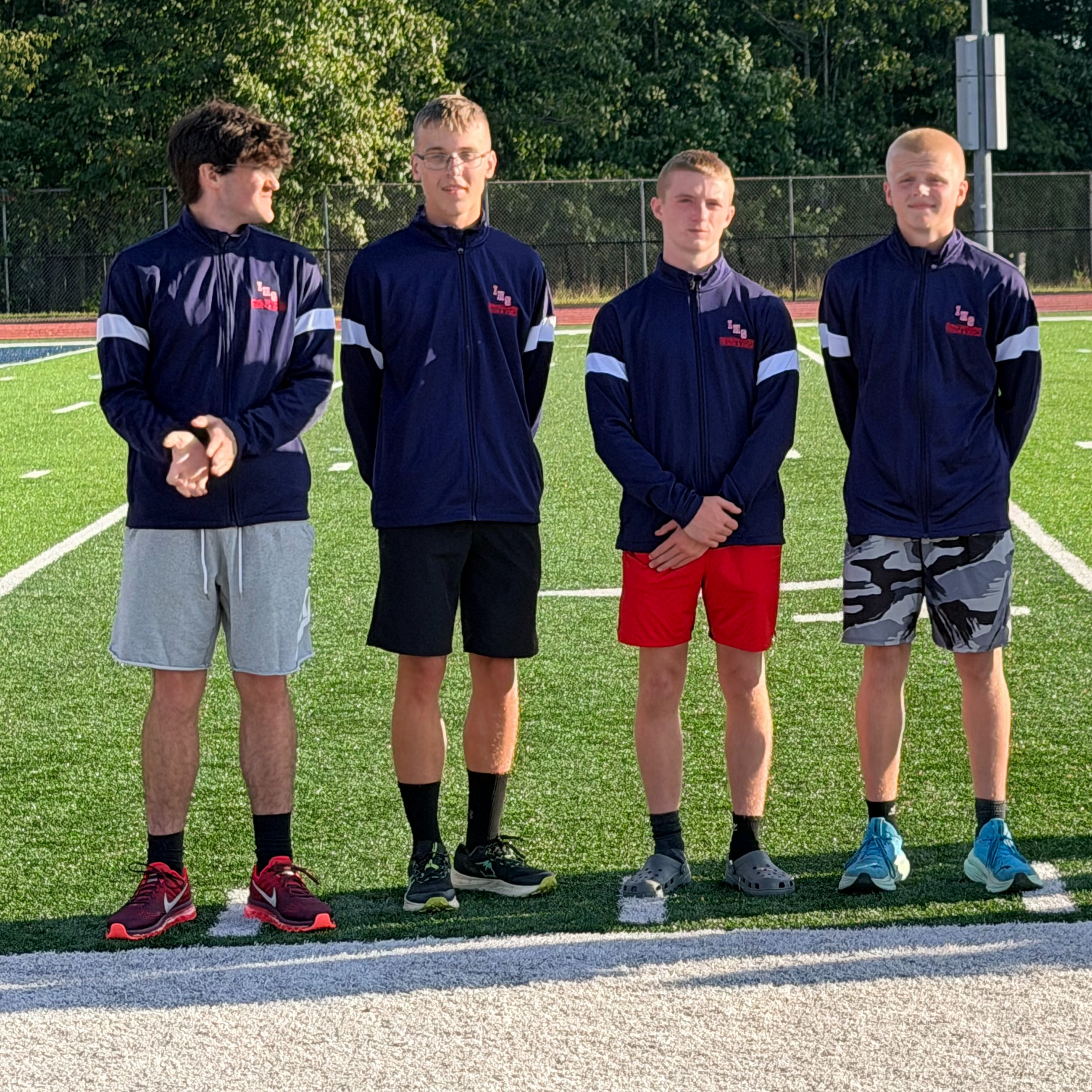Five people stand on a field, possibly for a team photo. Four are in athletic gear. One adult wears a hat.