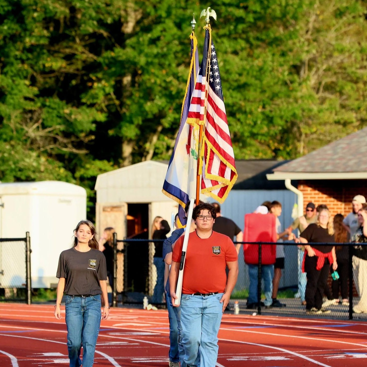 People walk on track; two carry flagpoles with American flag and other flags; spectators watch from sidelines.