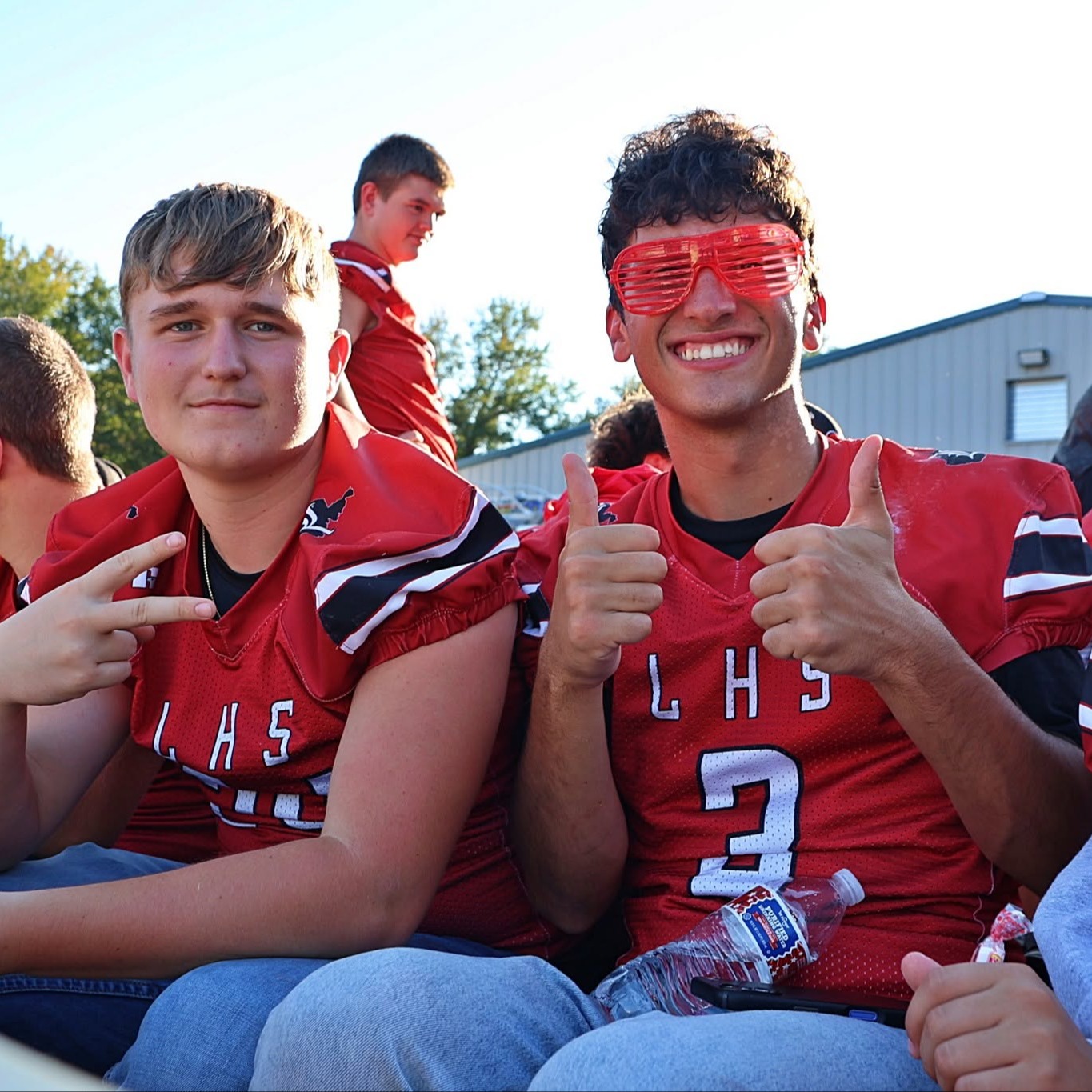 Group of students wearing red jerseys, seated in bleachers, one holding a water bottle.