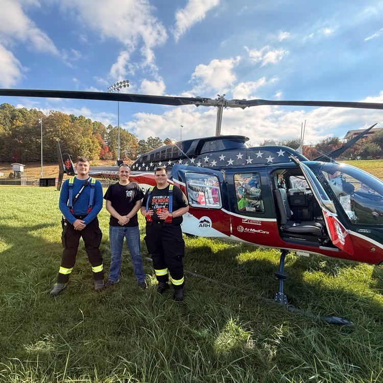 Three students stand in front of a red and white helicopter on a grassy field.