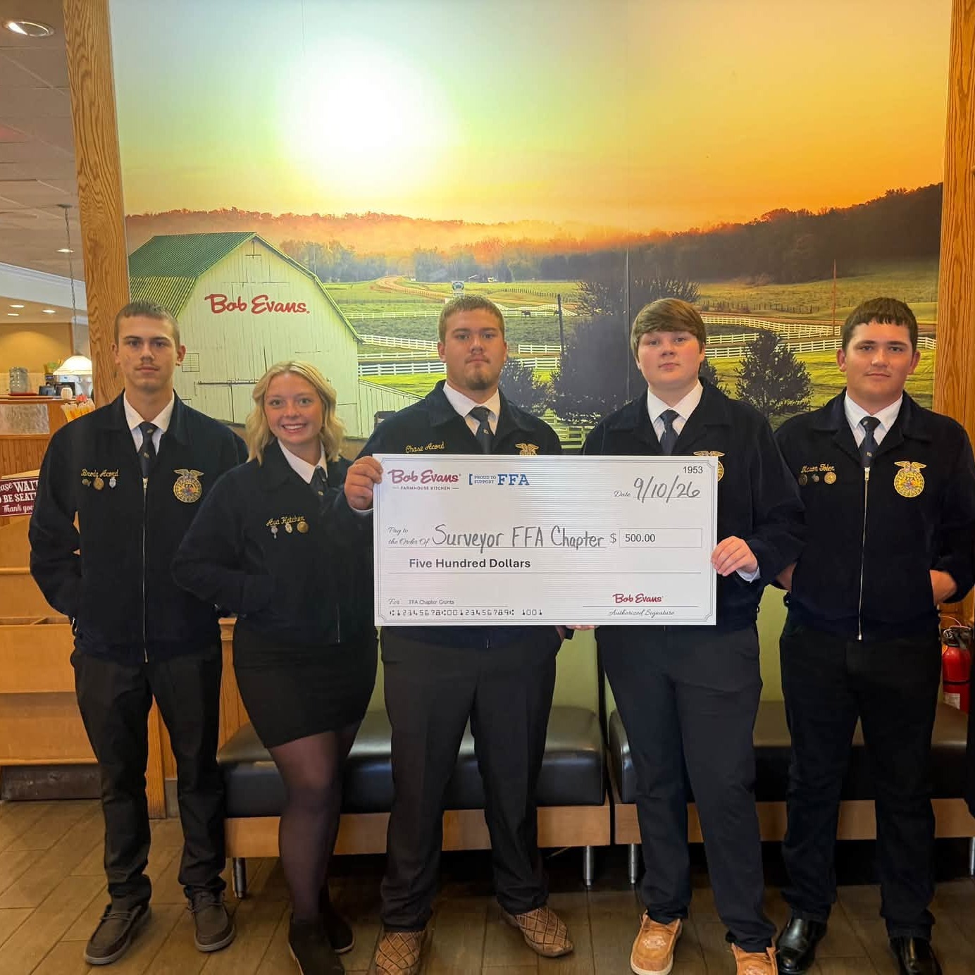 Five students and two adults pose holding a large check in front of a landscape mural.