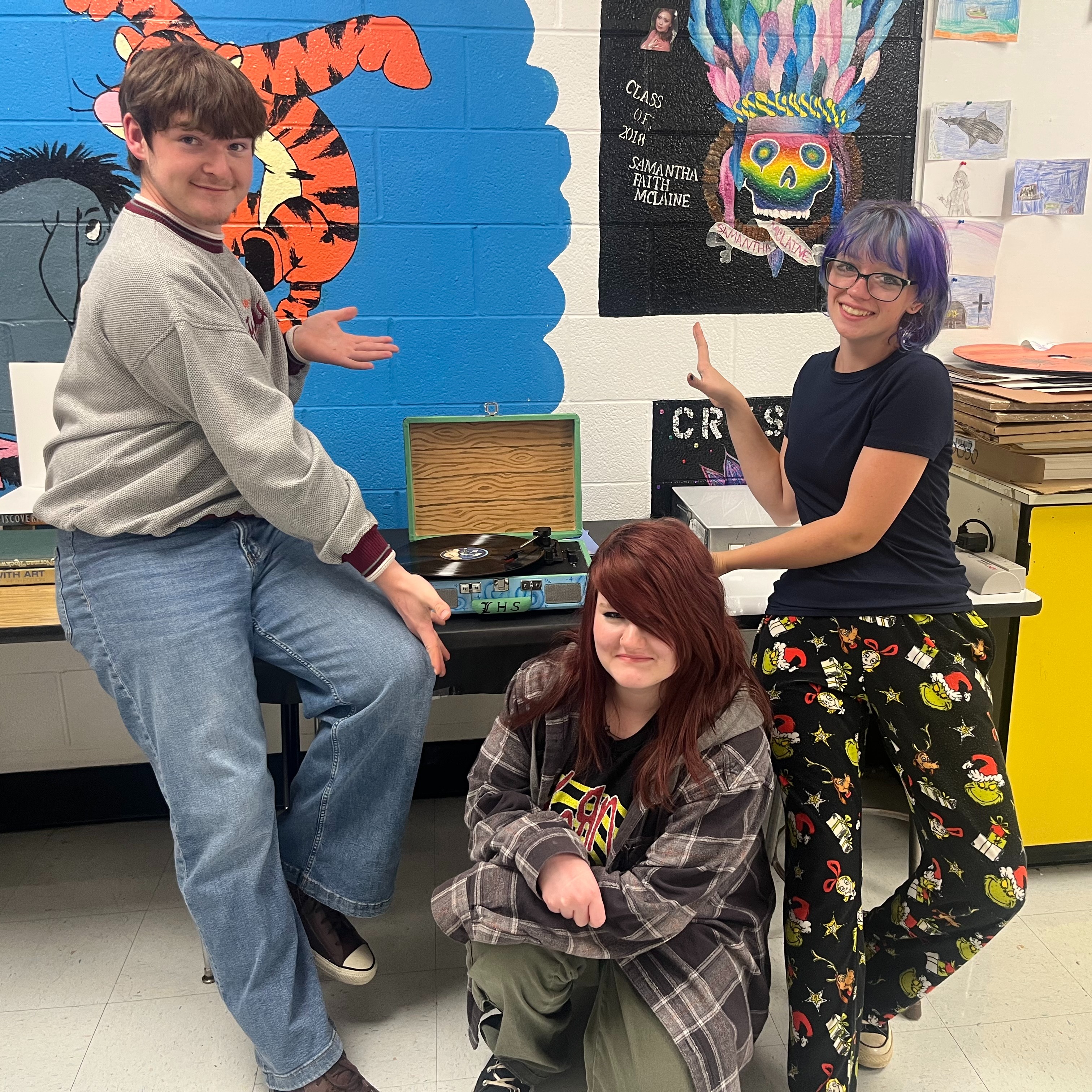 Three students pose for a picture with a colorful wall and boxes on a table.