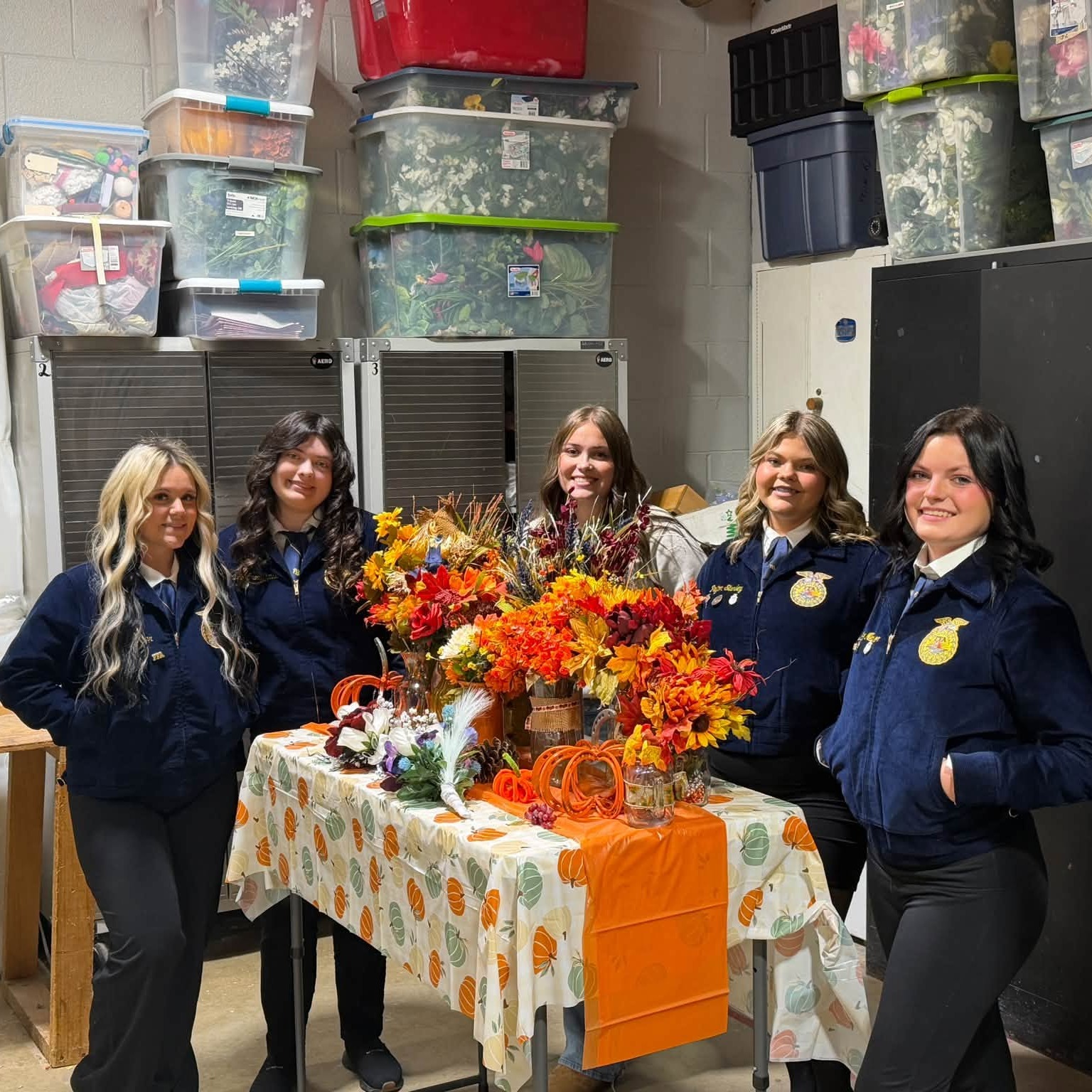 Five women in uniforms stand behind a table with an orange and yellow floral arrangement in front of a room with storage boxes.