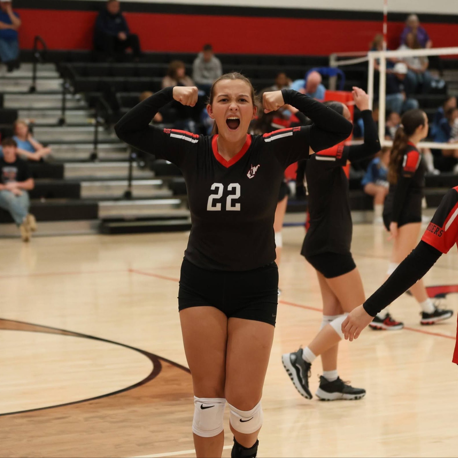 Volleyball players celebrate on court. One wears black with number 22. Another wears red with number 15.