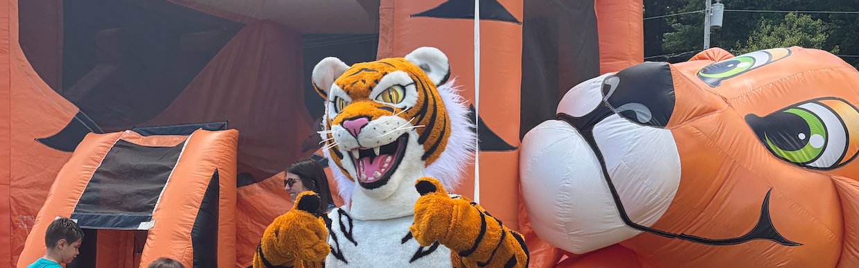 A tiger mascot stands on grass in front of a large orange tiger inflatable bounce house. Children play nearby.