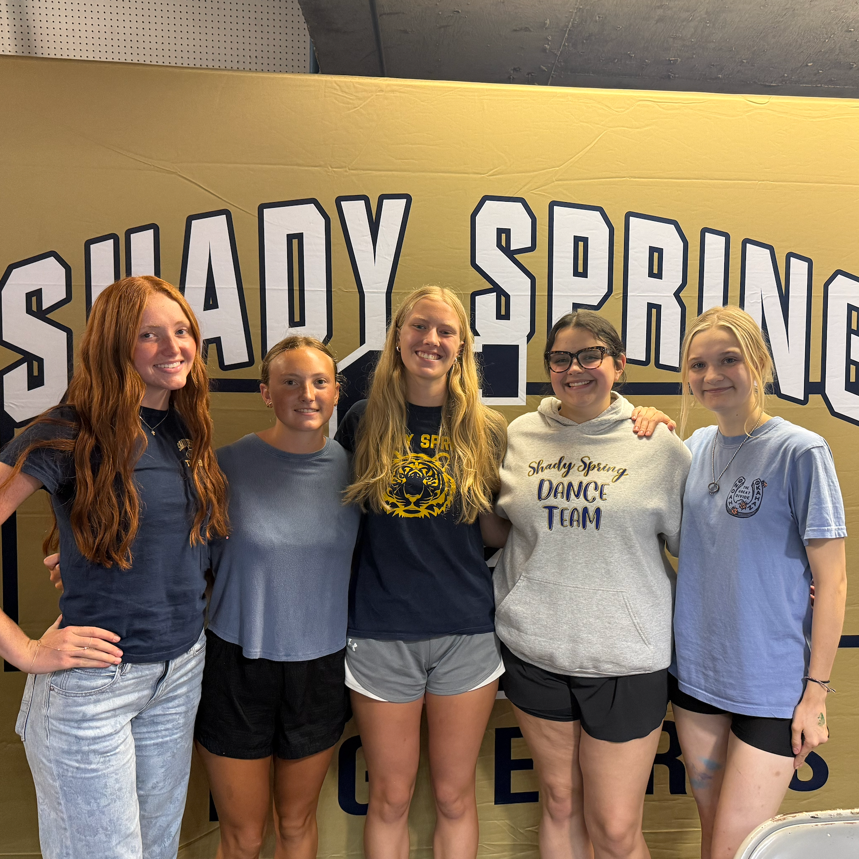 Five girls in blue shirts and black shorts pose for a photo in front of a gold backdrop.