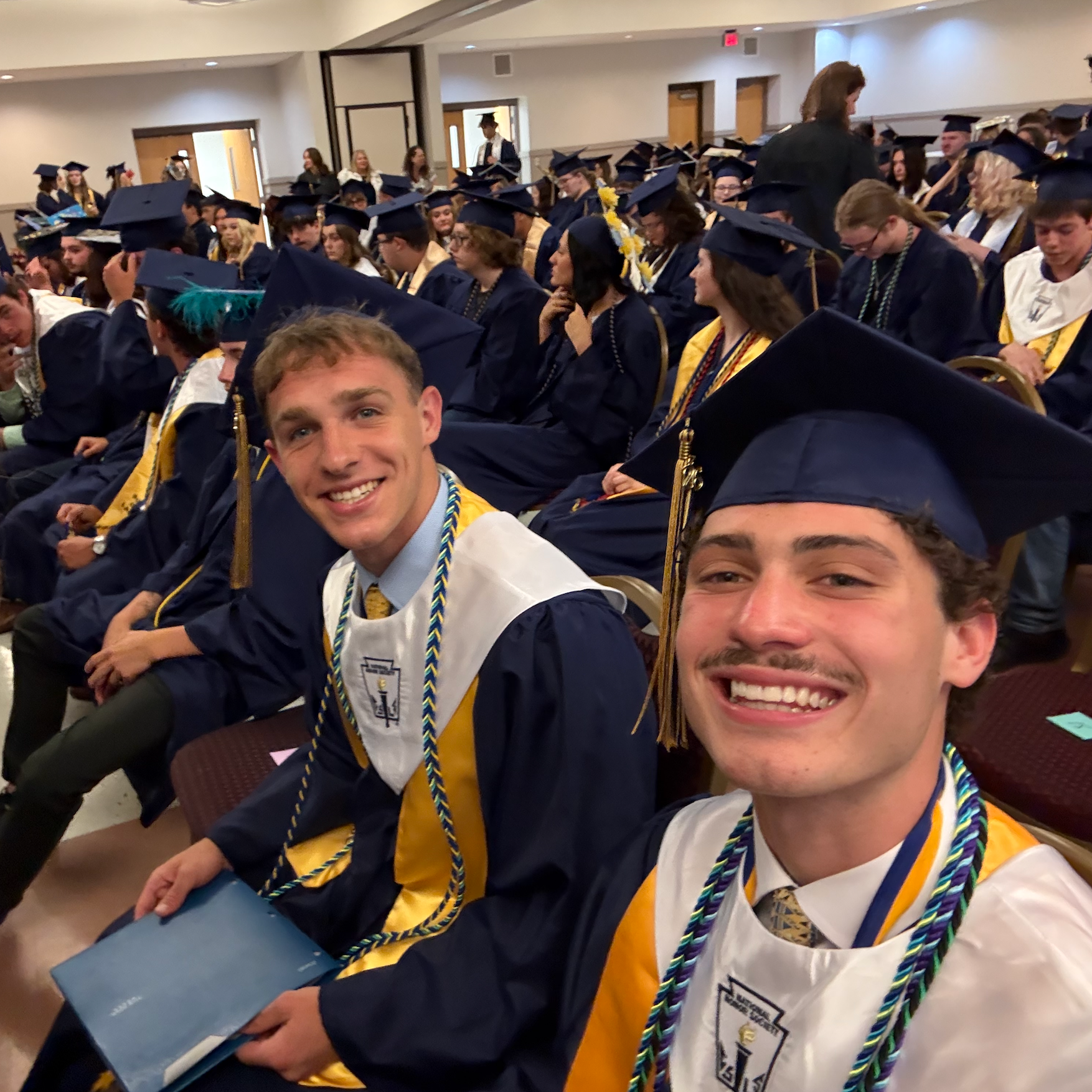 Graduates in caps and gowns sit among other graduates. Two men and a woman smile at the camera.