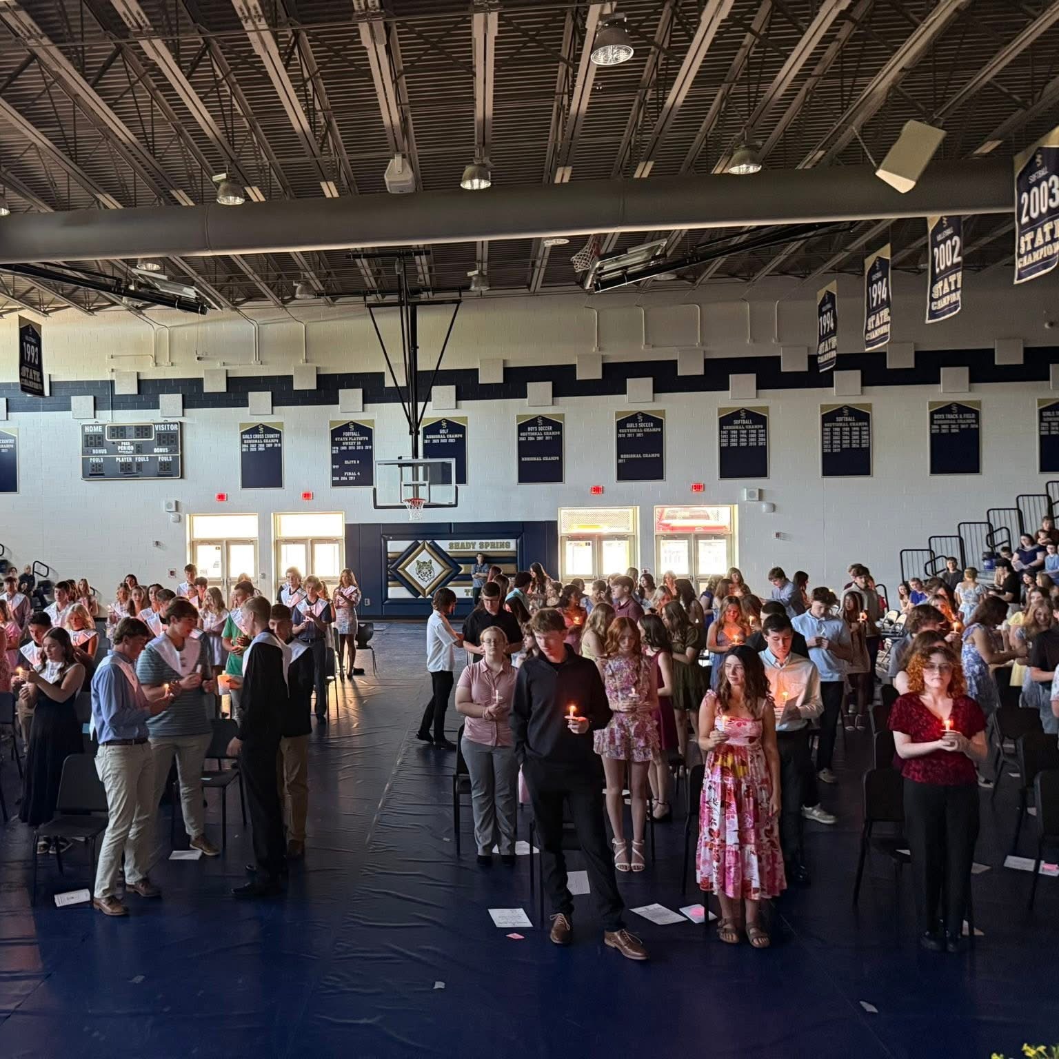 Indoor gymnasium with many people standing in a semi-circle, facing front, and a stage at the front.