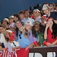 Students watching an outdoor event, some holding red cups, one holding a banner, sitting on bleachers.