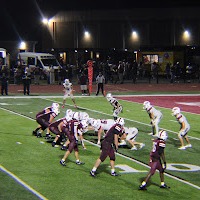 Night football game in progress; players in maroon uniforms, officials in black and white stripes, and spectators watching from stands.