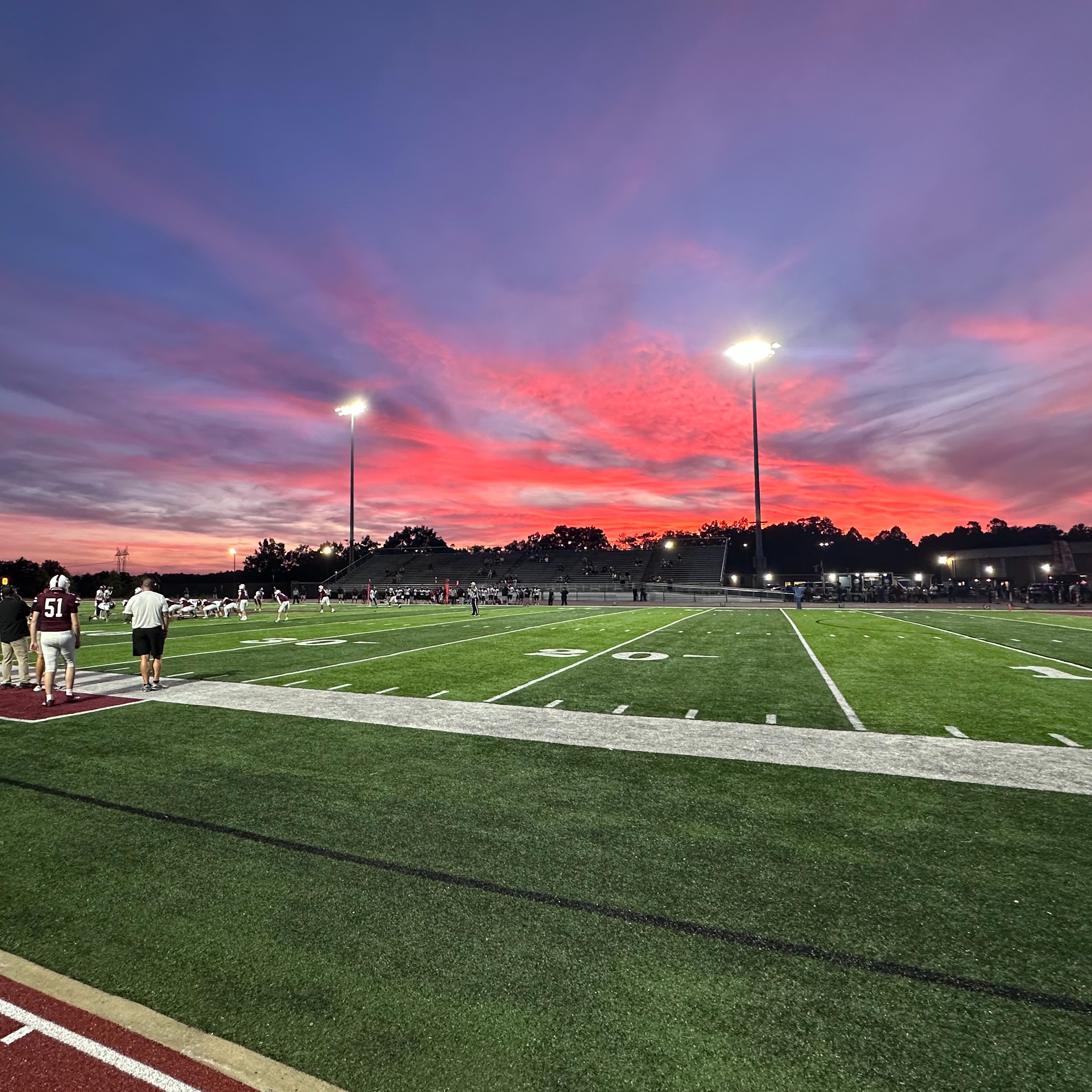 A football field with people standing on the sidelines and a colorful sunset sky with trees.