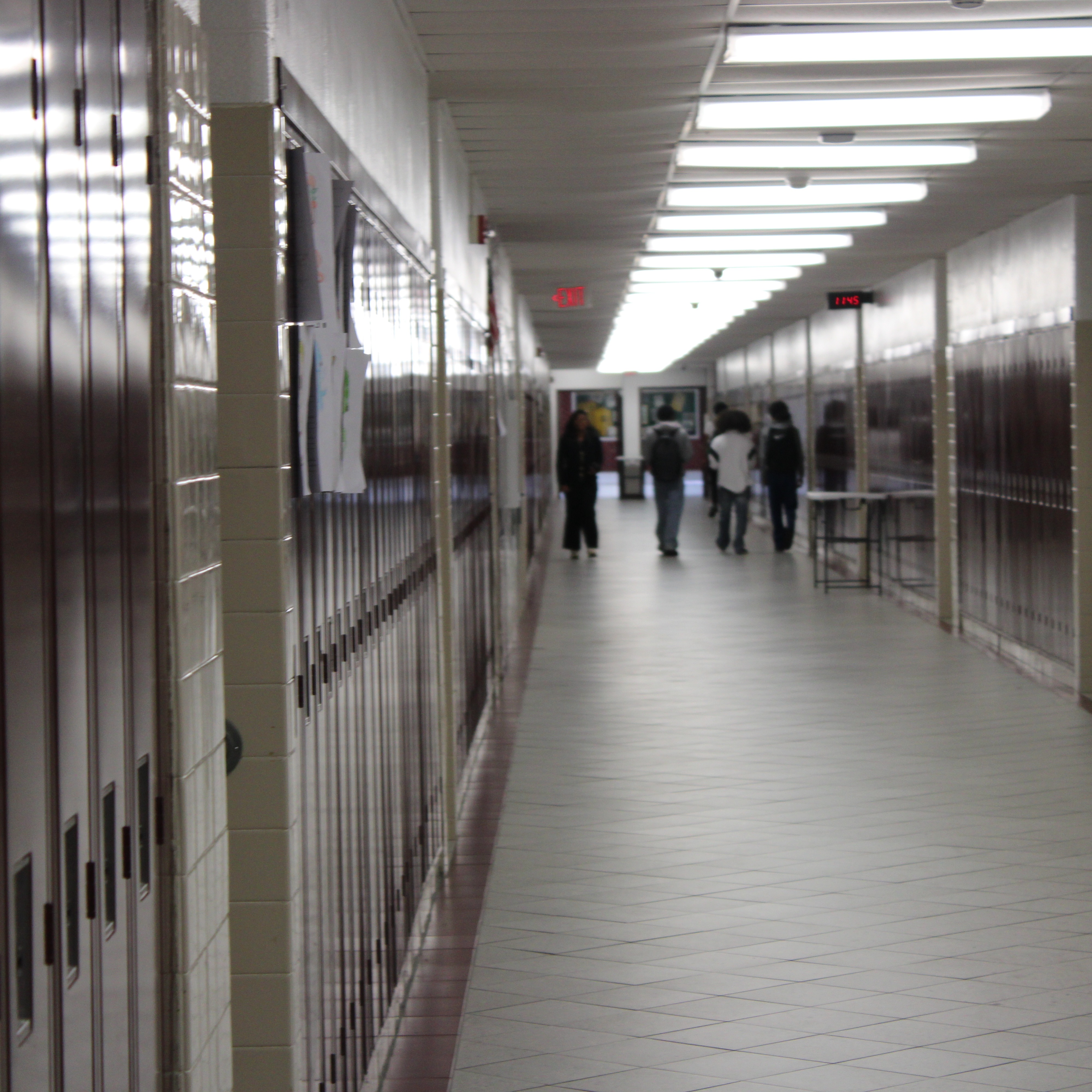 Long hallway with lockers on both sides. Several students walking. Papers attached to lockers.